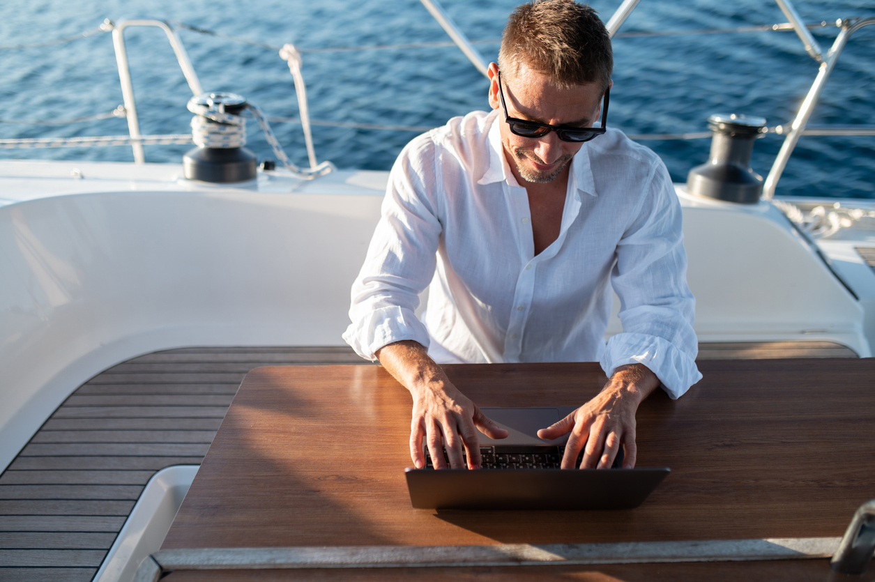 Work remotely. Man in white tshirt sitting on a yacht deck and typing on a laptop